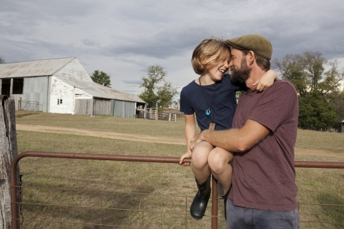 Happy girl sitting on gate hugging her father - Australian Stock Image