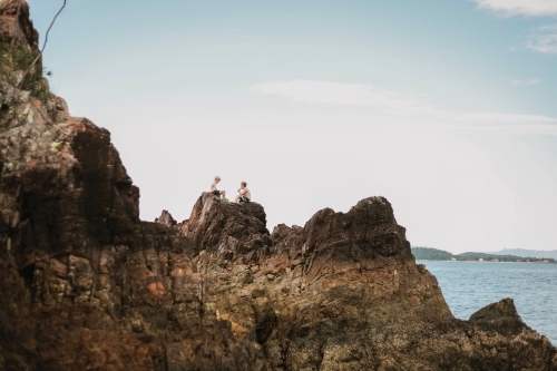 Kids sitting on rocks by the beach at Coral Beach near Shute Harbour in the Whitsundays - Australian Stock Image