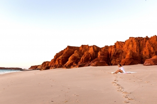 Landscape shot of a woman sitting on the sand with a tall red rock formation beside her - Australian Stock Image