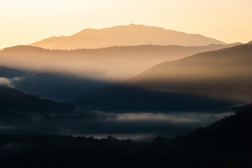 Layers of mountains on a cold foggy morning in winter with the early morning sun shining through - Australian Stock Image