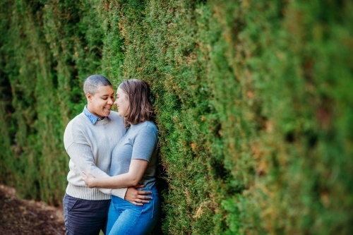 lgbtqi couple hugging each other and looking at each others faces while leaning on a wall with moss - Australian Stock Image