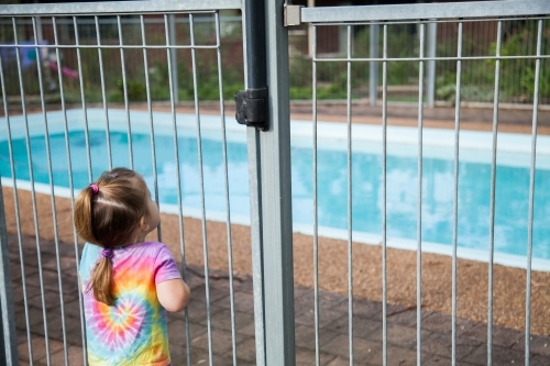 Little girl standing safely outside pool fence gate looking in - Australian Stock Image