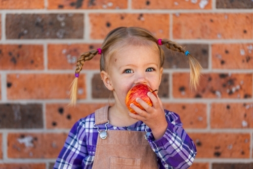 Little kid eating crunchy apple outside by brick wall of home - Australian Stock Image