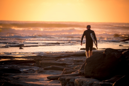 Lone surfer walking on rocks at sunrise - Australian Stock Image