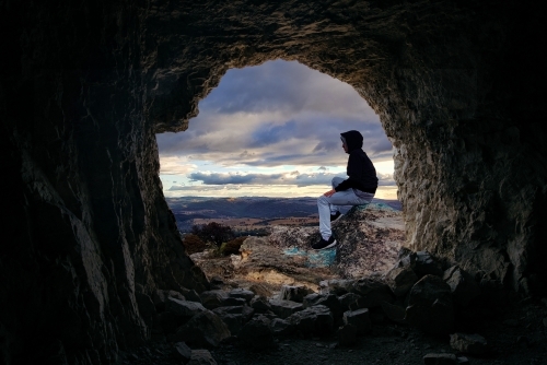 Looking through a cave to a male sitting on a rocky outcrop that looks over the valley - Australian Stock Image