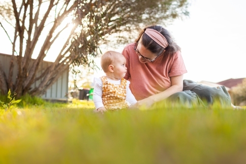 Low angle view of young mum and baby sitting on front lawn grass on spring afternoon - Australian Stock Image