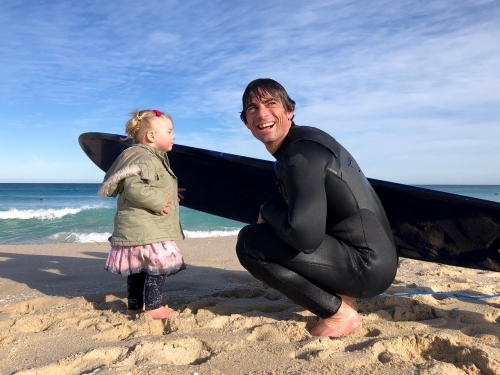 Male adult who’s just got out of surf kneeling down with female toddler on shoreline of beach - Australian Stock Image