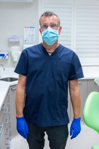 Male dentist wearing scrub top, gloves, mask and safety glasses in dental practice - Australian Stock Image