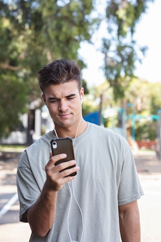 Male in his twenties wearing earphones and listening to music outdoors in the local park - Australian Stock Image