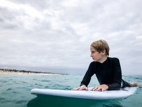 Male youth sitting on boogie board waiting for waves on overcast day - Australian Stock Image