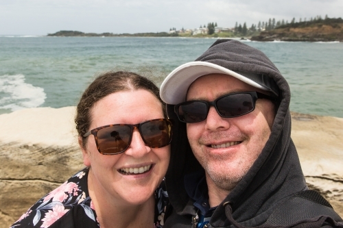 Man and woman selfie - husband and wife smiling wearing sunglasses at beach with ocean behind - Australian Stock Image