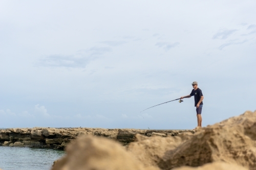 Man fishing from rocks with cloudy blue sky in the background and out of focus rocks in foreground - Australian Stock Image