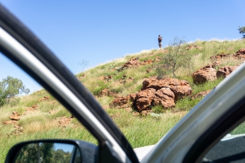 Man in distance standing on top of bushy rocky hill searching for service on his phone - Australian Stock Image