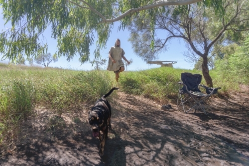 Man in hooded poncho towel walking down to riverside with dog leading the way, rugged bush of WA - Australian Stock Image