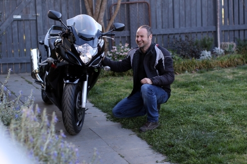 Man laughing and crouching next to motorbike in garden - Australian Stock Image