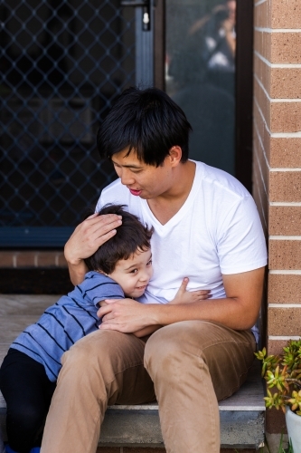 Man of Chinese ethnicity hugging mixed race son sitting on front steps of home