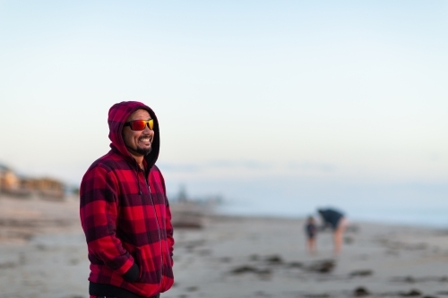 MAn on beach with hands in pockets with mother and child blurred in background - Australian Stock Image