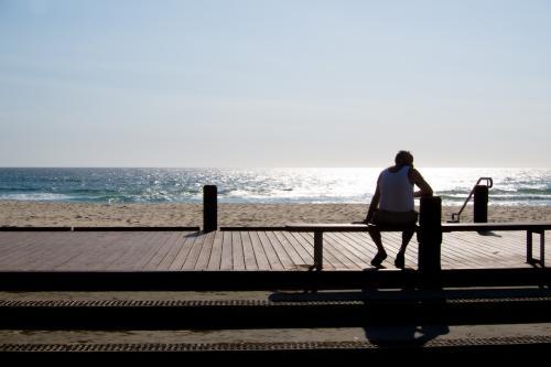 Man sitting on a bench silhouetted against the sun and sea - Australian Stock Image