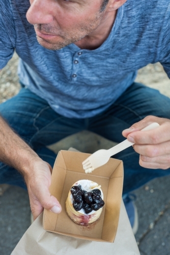 Man sitting on street curb eating bakery sweet - Australian Stock Image