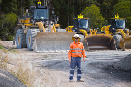 Man standing in the middle of the dirt path with payloaders at the back. - Australian Stock Image
