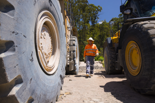 Man walking in between the massive wheels of payloader trucks on mine site - Australian Stock Image