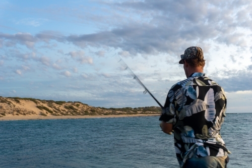 Man wearing floral shirt fishing from rocks with sunset sky in the background - Australian Stock Image