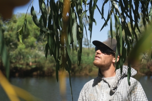 Man wearing hat and sunglasses looking through green foliage at sky, swimming hole in background - Australian Stock Image