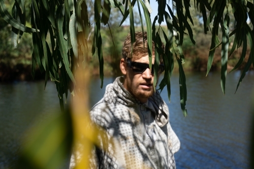 Man wearing sunglasses looking through green foliage at camera, swimming hole in background - Australian Stock Image