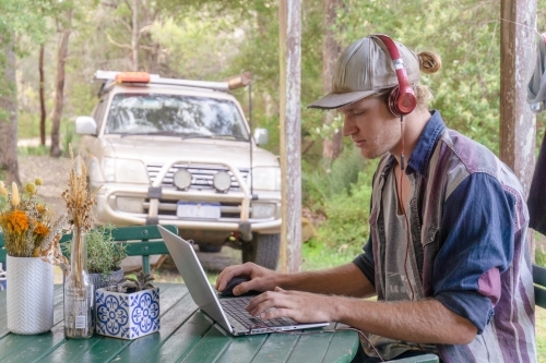 Man working remote on laptop with headphones on in the bush with trees and 4x4 car in background - Australian Stock Image