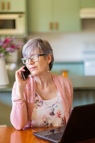 Mature woman on laptop at home using mobile phone to call for tech support - Australian Stock Image