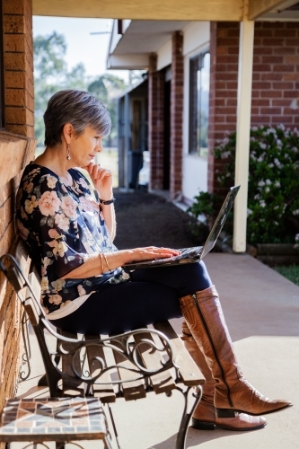 Mature woman sitting on park bench chair outside working on laptop in sunshine - Australian Stock Image