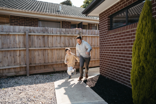 Middle-aged guy and young boy running on the pathway of their house. - Australian Stock Image