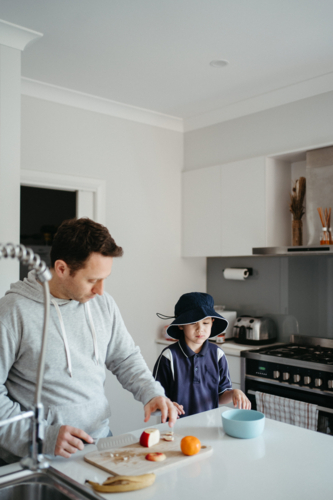 Middle-aged guy cutting fruit for school snacks on the kitchen counter with young boy - Australian Stock Image