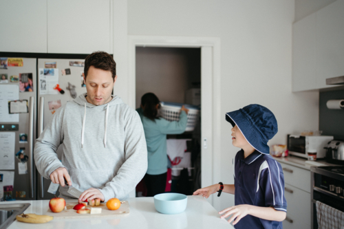 Middle-aged guy cutting fruit on the kitchen counter with young boy waiting beside him - Australian Stock Image