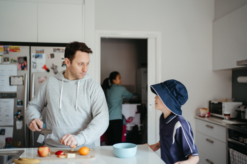 Middle-aged guy cutting fruit on the kitchen counter with young boy waiting beside him - Australian Stock Image