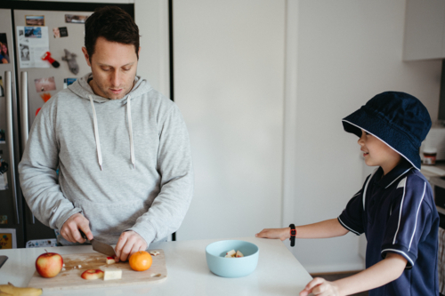 Middle-aged guy cutting fruits on the kitchen counter with young boy waiting beside him - Australian Stock Image