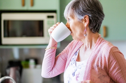 Middle aged woman in kitchen drinking mug of tea - Australian Stock Image