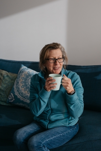 Middle aged woman sitting on couch, drinking hot beverage - Australian Stock Image