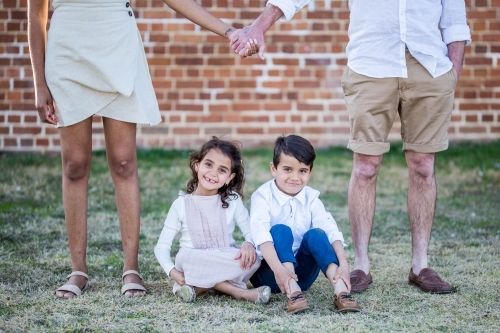 Mixed race aboriginal and caucasian twin children sitting on grass with parents holding hands - Australian Stock Image