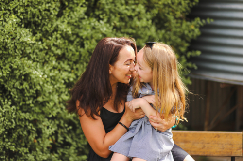 Mother and daughter snuggling in front of green hedge - Australian Stock Image