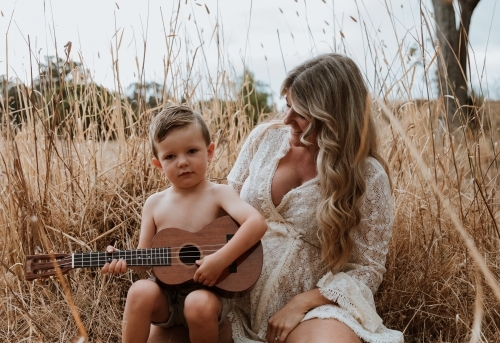 Mother and son sitting close together outside in field with small guitar - Australian Stock Image