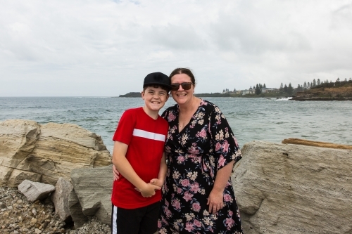 Mother and son standing together smiling on rock wall in front of beach water - Australian Stock Image