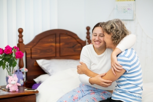 Mother and teen daughter sitting on bed and laughing with arms around each other - Australian Stock Image