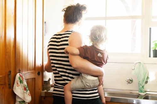 Mother at sink in the kitchen holding baby on hip - Australian Stock Image