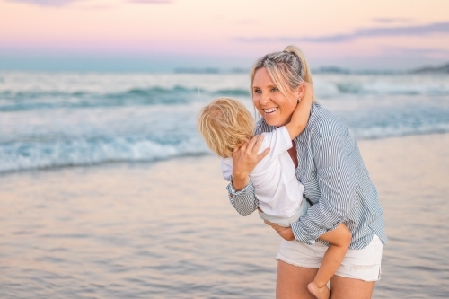 Mother carrying son on the beach in pink twilight light. Sunset on the Gold Coast. - Australian Stock Image