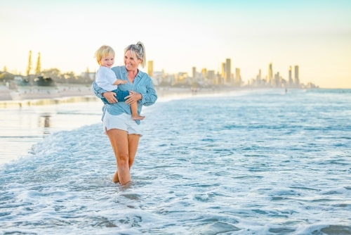 Mother carrying son on the beach with Gold Coast city skyline in background - Australian Stock Image