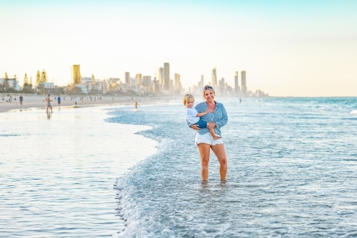 Mother carrying son on the beach with Gold Coast city skyline in background - Australian Stock Image