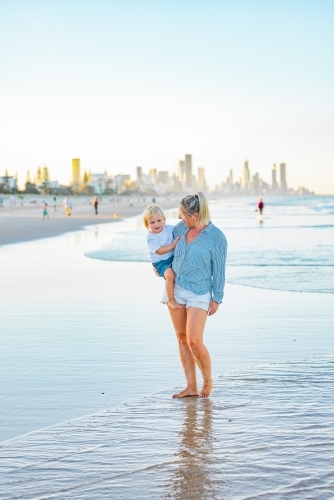 Mother carrying son on the beach with Gold Coast city skyline in background - Australian Stock Image