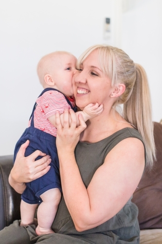 Mother holding baby boy smiling while he kisses her cheek - Australian Stock Image