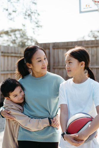 Mother hugs her two young children outside in their backyard. - Australian Stock Image